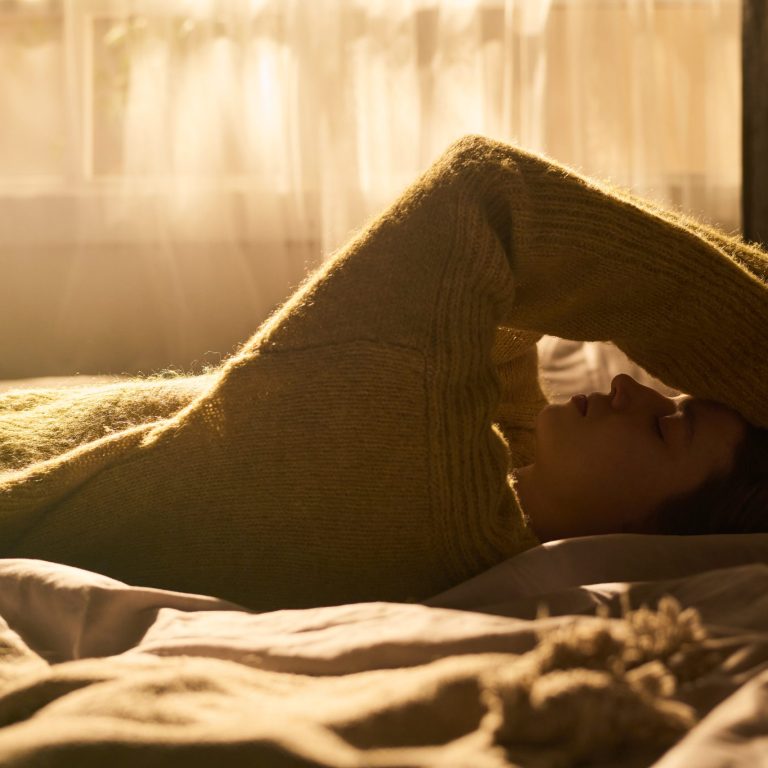 Young woman lying on bed resting with arm covering forehead, eyes closed, appearing relaxed in softly lit bedroom with sunlight streaming through window